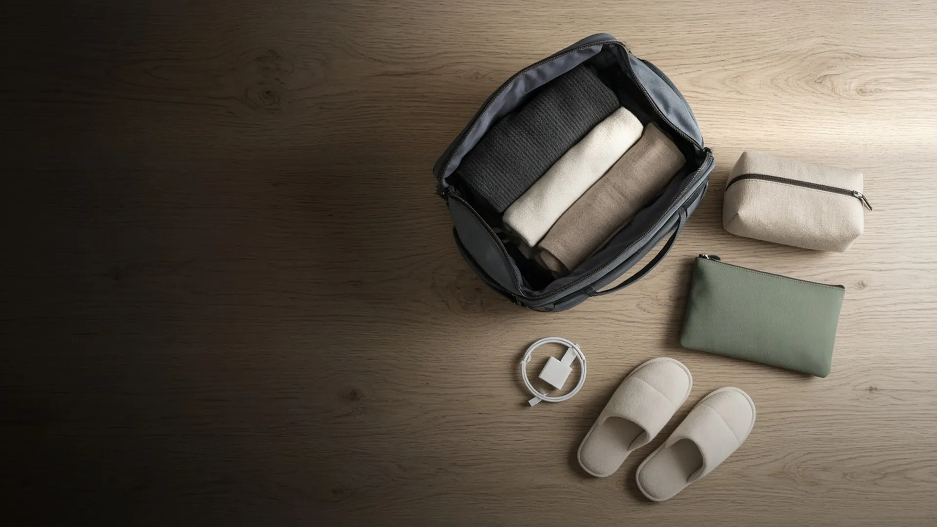 An open bag on an oak table with clothes, pouches, a charger, and slippers