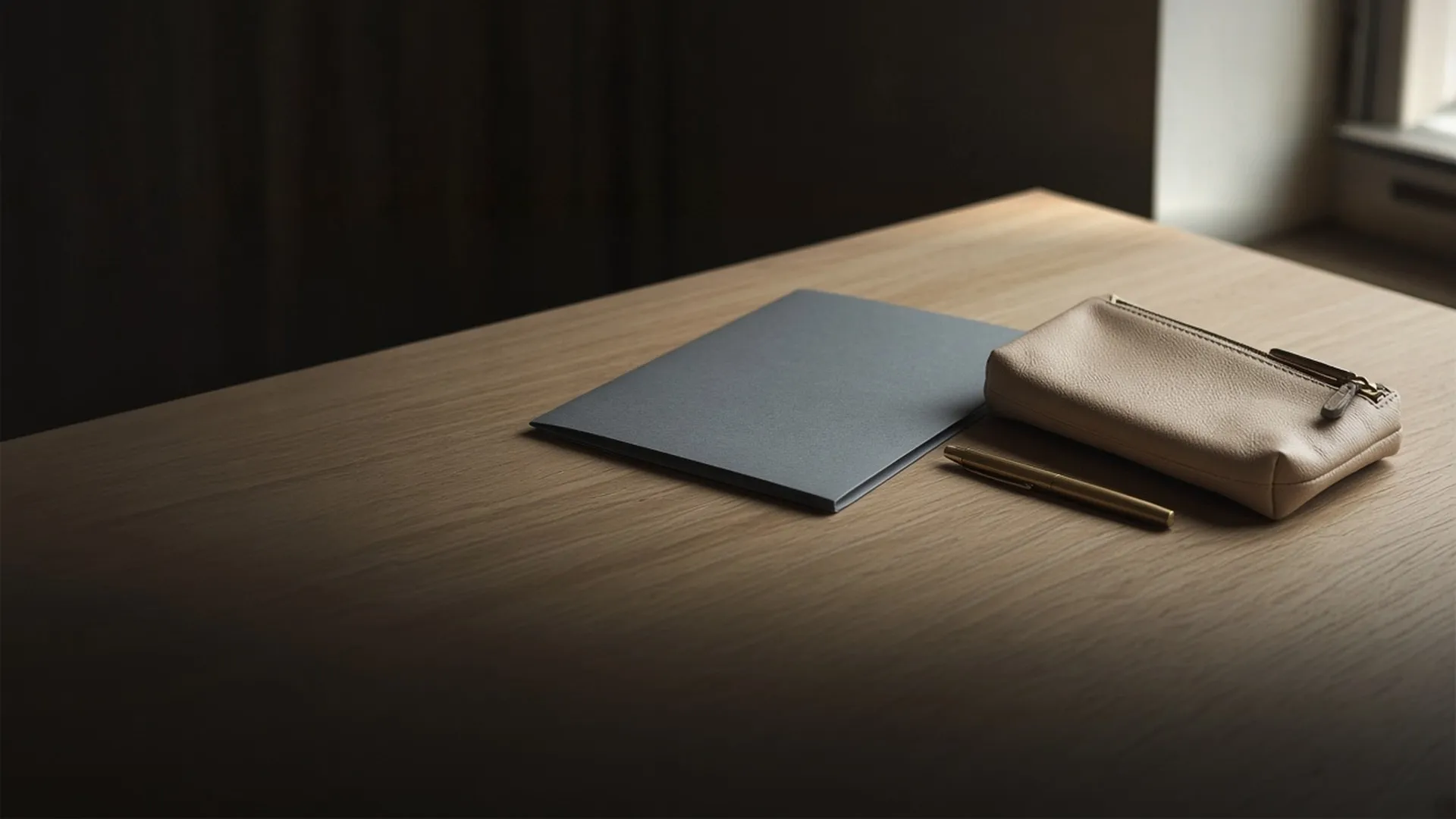 A closed document folder and a zip pouch beside a pen on an oak desk